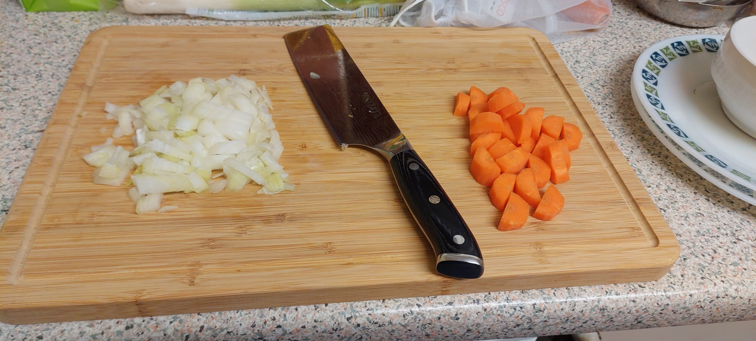 A chopping board with a pile of finely dinced onions on the left a pile of diced carrots on the right and a large chopping knife in between.